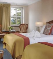 Hotel room with two beds, gold and red bedcovers, a table with two chairs, a window with closed curtains, and a dressing gown on the bed.