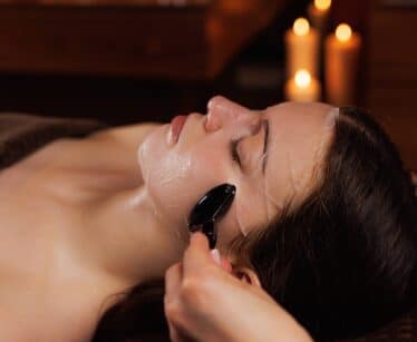 A person receives a facial massage with a jade roller while lying down; background features lit candles.
