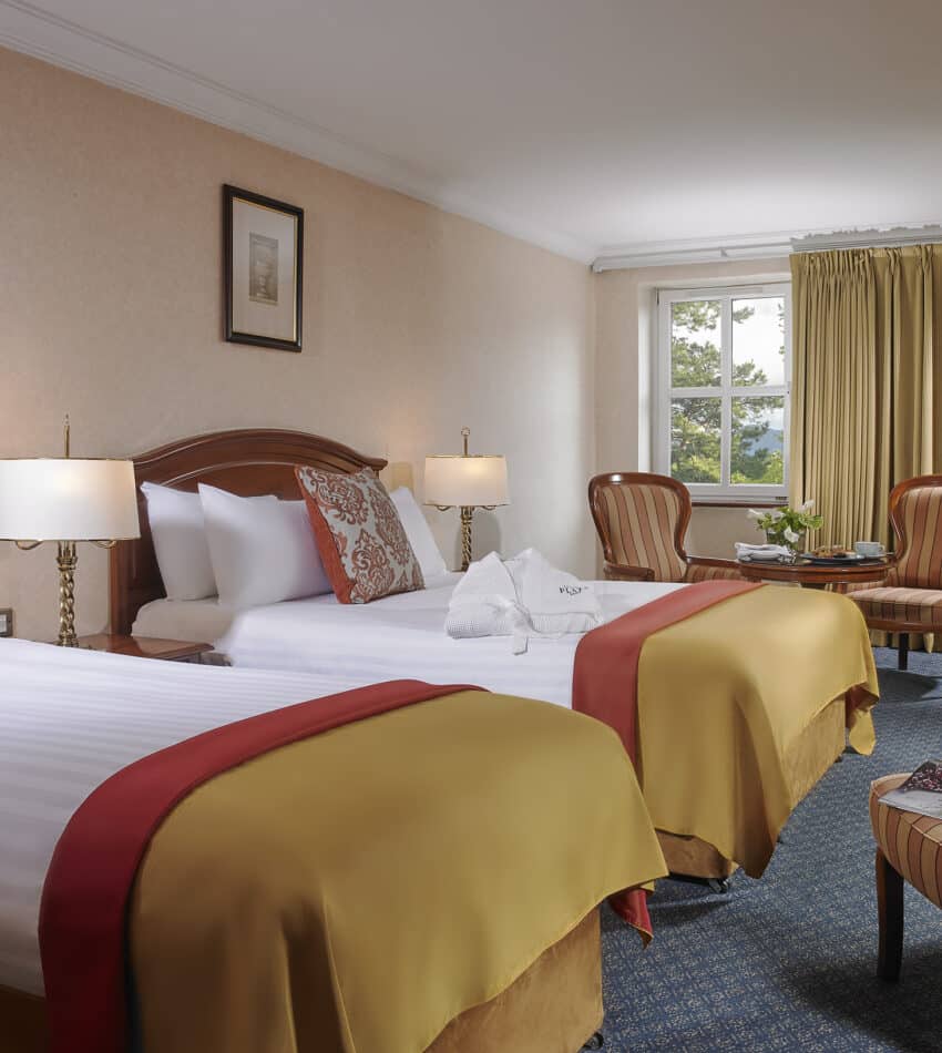 Hotel room with two double beds, gold and red bedding, armchairs by a window, lamps, a desk, and framed art on light-coloured walls.
