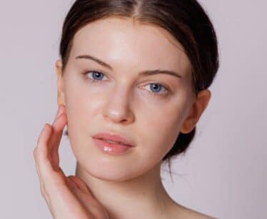 A woman with fair skin and blue eyes touches her face with one hand, looking directly at the camera against a plain light background.