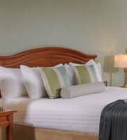 A neatly made bed with white linen, striped pillows, two bedside tables with lamps, and a framed picture on a light-coloured wall.
