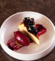 A slice of cheesecake topped with berry compote, served with a scoop of berry sorbet and sauce on a white plate.