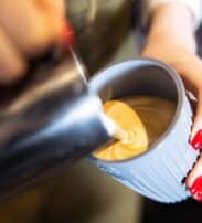 Close-up of a person pouring steamed milk from a metal jug into a cup of coffee, creating latte art.