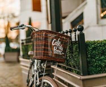 A bicycle with a wicker basket labelled "Café du Parc" is parked next to a metal railing and neatly trimmed greenery outside a building.