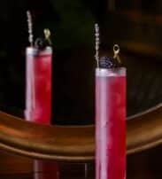 Tall glass of pink cocktail with ice, garnished with a blackberry and a sprig, placed on a dark surface in front of a mirror.