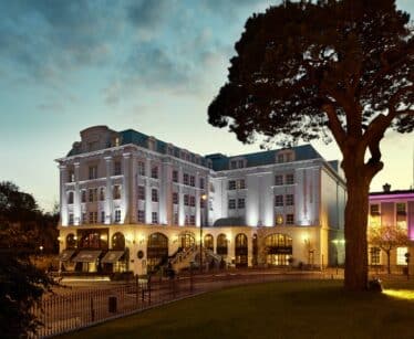A large, elegant hotel building with illuminated exterior lights at dusk, surrounded by trees and featuring several arched windows and entrances.