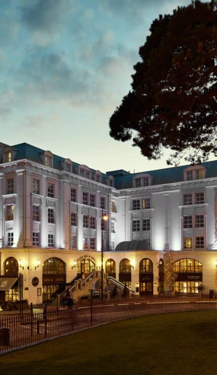 A large, elegant hotel building with illuminated exterior lights at dusk, surrounded by trees and featuring several arched windows and entrances.