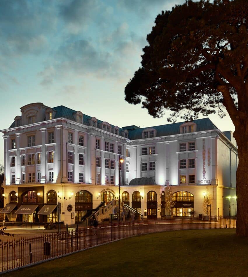 A large, elegant hotel building with illuminated exterior lights at dusk, surrounded by trees and featuring several arched windows and entrances.