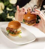 A person with painted nails holds a slider burger above a plate with another burger; both have lettuce and lightly toasted baps.