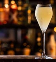 A cocktail in a tall, stemmed glass sits on a bar counter with a blurred background of bottles and warm lights.