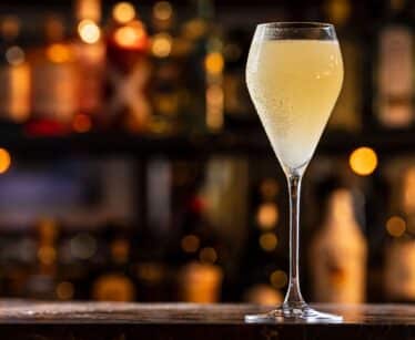 A cocktail in a tall, stemmed glass sits on a bar counter with a blurred background of bottles and warm lights.