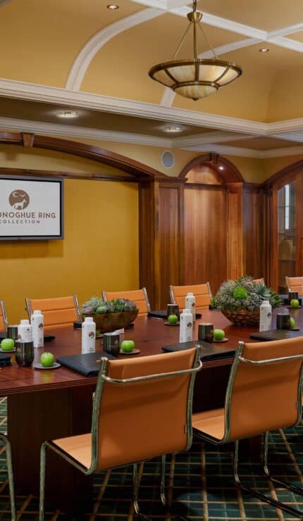 A conference room with a long table, tan chairs, notepads, bottled water, small plants, and a wall-mounted screen displaying "O'Donoghue Ring Collection.