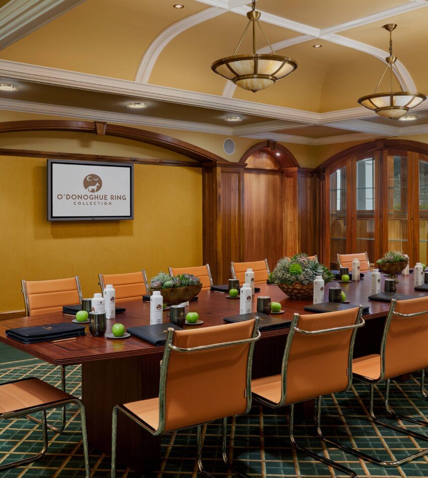 A conference room with a long table, tan chairs, notepads, bottled water, small plants, and a wall-mounted screen displaying "O'Donoghue Ring Collection.