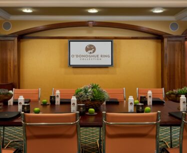 Conference room with beige chairs, a table set with bottled water, apples, notebooks, and succulent centrepieces, and a screen displaying "O'Donoghue Ring Collection.