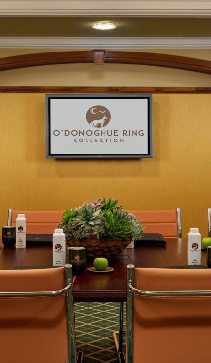 Conference room with beige chairs, a table set with bottled water, apples, notebooks, and succulent centrepieces, and a screen displaying "O'Donoghue Ring Collection.