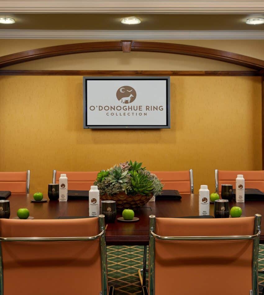 Conference room with beige chairs, a table set with bottled water, apples, notebooks, and succulent centrepieces, and a screen displaying "O'Donoghue Ring Collection.