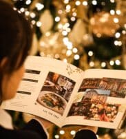 Person reading a magazine featuring food and restaurant images in front of a decorated, lit Christmas tree.