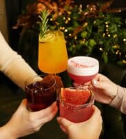 Four hands holding assorted colourful cocktails together in a toast, with greenery and festive lights in the background.