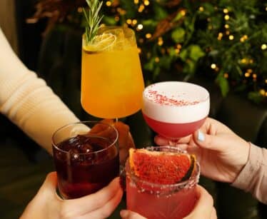 Four hands holding assorted colourful cocktails together in a toast, with greenery and festive lights in the background.