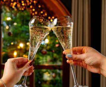 Two hands clink champagne glasses in front of a window with festive holiday lights and a decorated Christmas tree in the background.