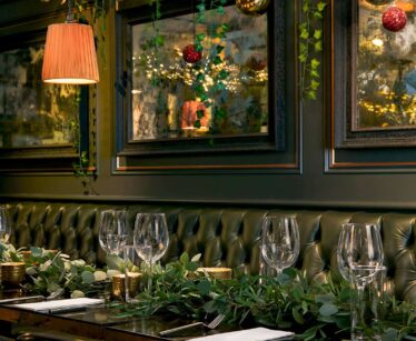 A restaurant table set with wine glasses and cutlery, decorated with greenery; festive baubles and lights hang above, with framed art on a dark green wall.