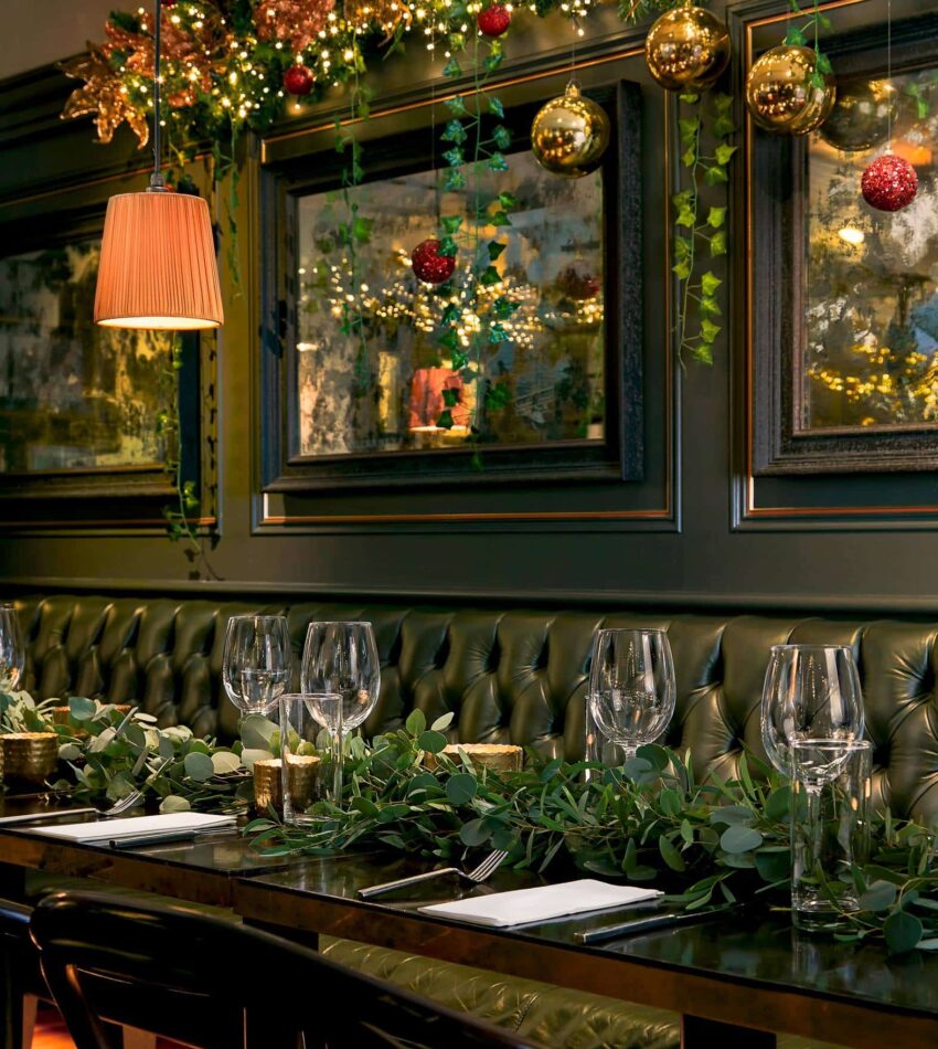 A restaurant table set with wine glasses and cutlery, decorated with greenery; festive baubles and lights hang above, with framed art on a dark green wall.