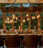 A dining table laid for a meal with brown chairs, festive decorations, candles, and hanging lights, in front of large windows with greenery and ornaments outside.