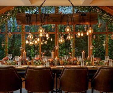 A dining table laid for a meal with brown chairs, festive decorations, candles, and hanging lights, in front of large windows with greenery and ornaments outside.