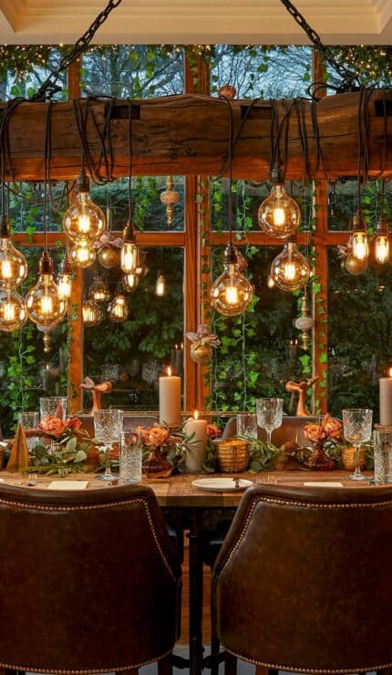 A dining table laid for a meal with brown chairs, festive decorations, candles, and hanging lights, in front of large windows with greenery and ornaments outside.