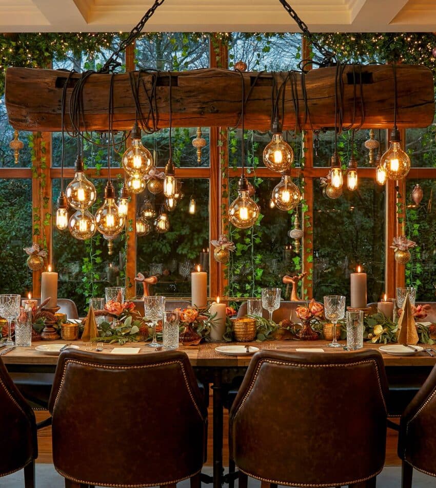 A dining table laid for a meal with brown chairs, festive decorations, candles, and hanging lights, in front of large windows with greenery and ornaments outside.