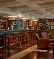 A decorated bar lounge with leather chairs, a wooden bar, festive garlands, and a lit Christmas tree near the window. Framed pictures hang on the walls.
