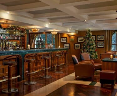 A decorated bar lounge with leather chairs, a wooden bar, festive garlands, and a lit Christmas tree near the window. Framed pictures hang on the walls.