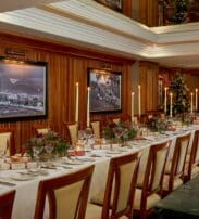 A long dining table set for a formal meal with elegant crockery, floral centrepieces, Christmas decorations, and framed black-and-white photos on wood-panelled walls.