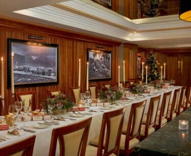 A long dining table set for a formal meal with elegant crockery, floral centrepieces, Christmas decorations, and framed black-and-white photos on wood-panelled walls.