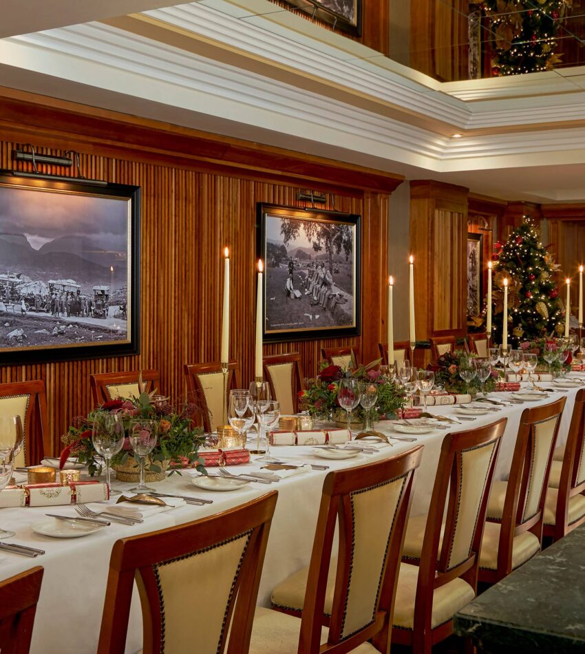 A long dining table set for a formal meal with elegant crockery, floral centrepieces, Christmas decorations, and framed black-and-white photos on wood-panelled walls.
