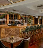 A barman stands behind a marble-topped bar with green stools in an elegant, warmly lit lounge decorated with greenery and plants.
