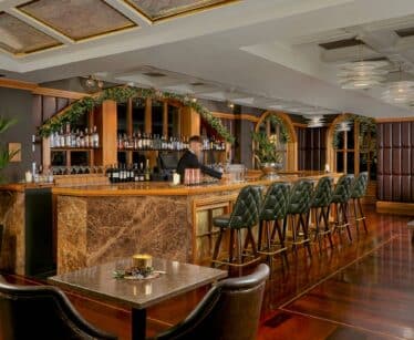 A barman stands behind a marble-topped bar with green stools in an elegant, warmly lit lounge decorated with greenery and plants.