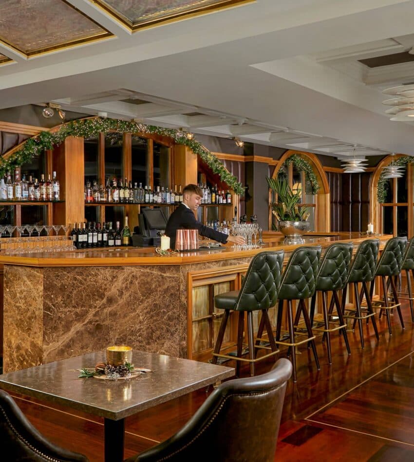 A barman stands behind a marble-topped bar with green stools in an elegant, warmly lit lounge decorated with greenery and plants.