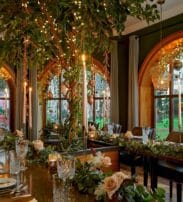 Elegant dining room with large arched windows, a long table set with glassware and floral arrangements, and fairy lights decorating a central tree and ceiling.