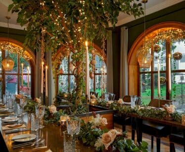 Elegant dining room with large arched windows, a long table set with glassware and floral arrangements, and fairy lights decorating a central tree and ceiling.
