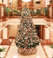 A large, decorated Christmas tree stands in the centre of an ornate, indoor lobby with marble accents and holiday greenery on the railings.