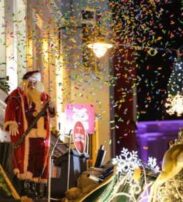 A person dressed as Father Christmas stands on a decorated float with a reindeer figure, surrounded by falling confetti and illuminated festive decorations at night.