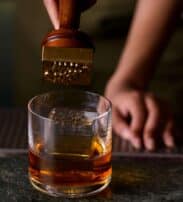 A person presses a metal stamp onto a large ice cube in a glass of whisky on a bar countertop.