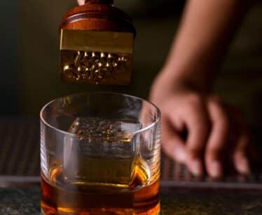 A person presses a metal stamp onto a large ice cube in a glass of whisky on a bar countertop.