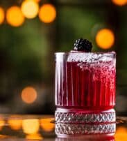 A dark red cocktail in a glass with a salted rim, garnished with a blackberry, sits on a reflective surface with blurred warm lights in the background.