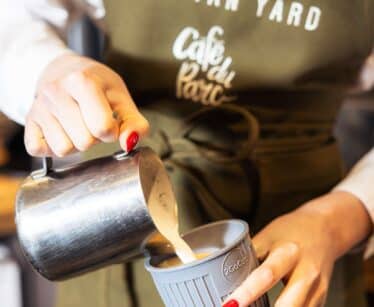 A person wearing an apron labelled "The Tan Yard" pours steamed milk from a metal jug into a reusable cup.