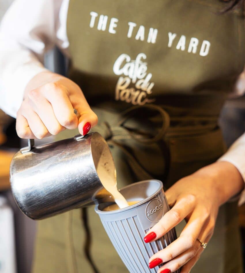 A person wearing an apron labelled "The Tan Yard" pours steamed milk from a metal jug into a reusable cup.