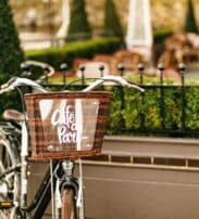 A black bicycle with a brown wicker basket labelled "Café à Paris" is parked by a fenced outdoor seating area with greenery.