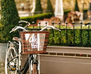 A black bicycle with a brown wicker basket labelled "Café à Paris" is parked by a fenced outdoor seating area with greenery.