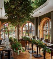 Elegant dining room with large arched windows, long wooden tables decorated with greenery, brown leather chairs, hanging lights, and indoor plants, including a tall central tree.